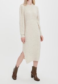 Kvinna klädd i en beige ribbad stickad midiklänning med sidofålla och långa puffärmar, tillsammans med bruna texturerade ankelskors med blockklackar.