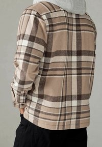 Beżowa i brązowa koszula flanelowa w kratę, z długimi rękawami, o miękkiej strukturze, z zapięciem na guziki przy mankietach i zaokrąglonym dołem, noszona na szarym topie z kapturem.