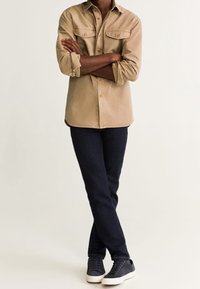 Persona de pie con los brazos cruzados, vistiendo una camisa beige abotonada, jeans azul oscuro y zapatillas navy con suelas blancas sobre un fondo claro.