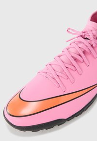 Scarpa da calcio Nike con una tomaia liscia di colore rosa, dettagli neri, un design Swoosh arancione e lacci piatti rosa. Suola nera testurizzata.