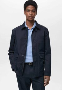 Giacca blu navy in misto cotone con tasche frontali, chiusura a bottoni e colletto morbido, abbinata a una polo testa di moro in tessuto leggero.