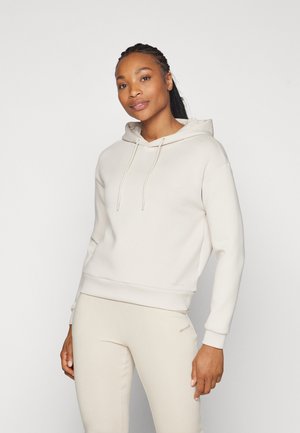 Femme aux cheveux tressés portant un sweat à capuche beige clair et un pantalon assorti, debout devant un fond blanc uni avec une expression détendue.