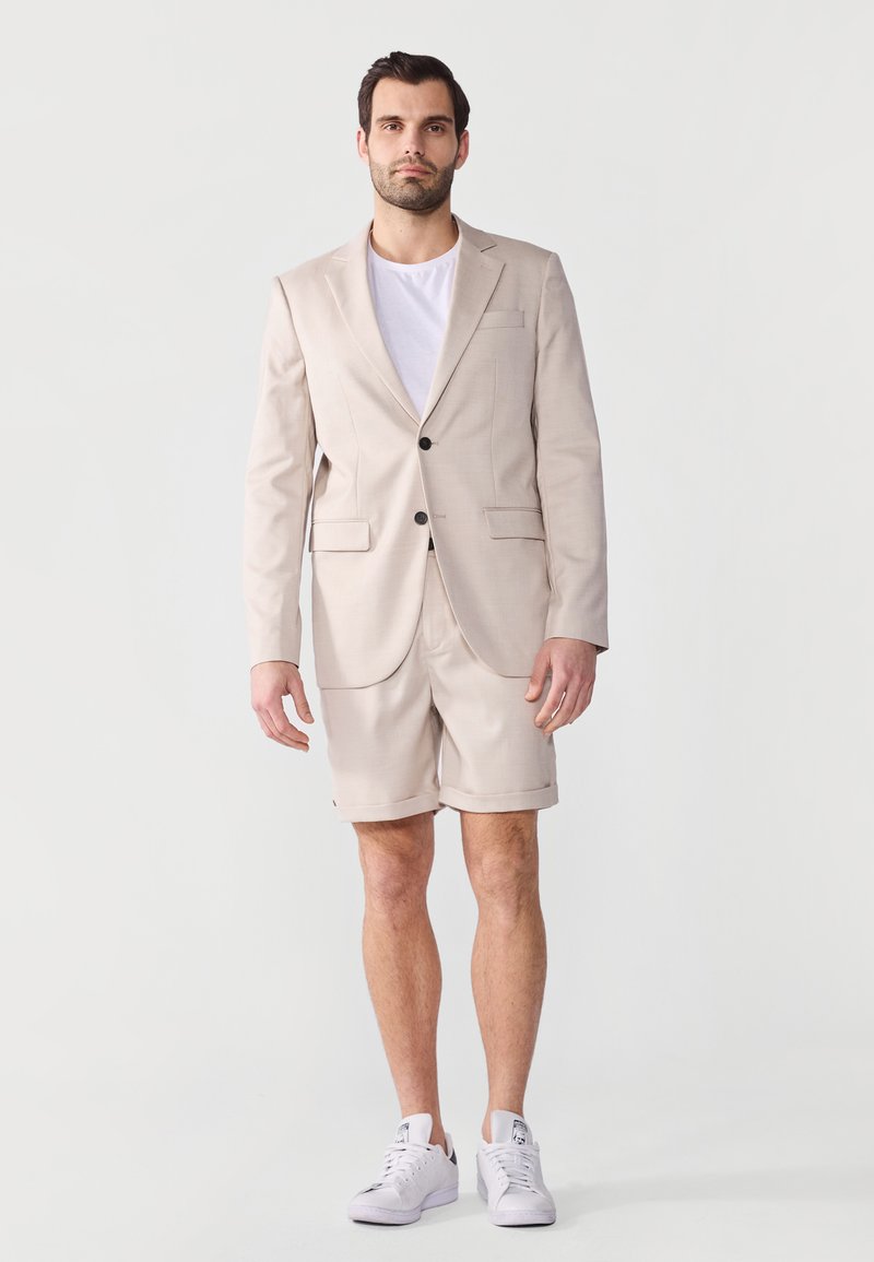 Veste de costume beige avec deux boutons, design ajusté et revers crantés, associée à un short assorti. Portée sur un t-shirt blanc et des baskets blanches.