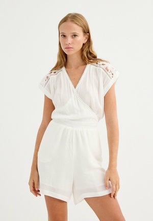 Mujer joven con un mono corto blanco de manga corta, con hombros de encaje y delantero cruzado, posando frente a un fondo claro y liso.