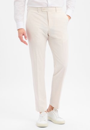 Mann trägt beige maßgeschneiderte Hose, weißes Hemd und weiße Sneakers, steht mit einer Hand in der Tasche vor weißem Hintergrund.