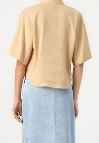 Personne portant une chemise beige à manches courtes avec col et une jupe en jean bleu clair taille haute, vue de dos.