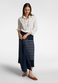 Camicia bianca a bottoni, gonna plissettata blu navy con righe bianche, scarpe basse marroni, gi jewelry a strati, mentre tiene una giacca blu navy.