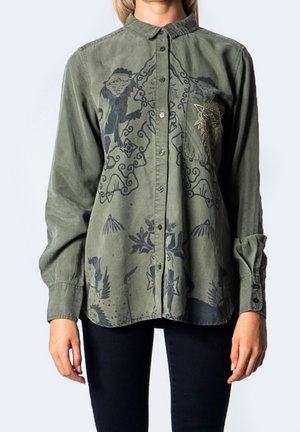 Długa koszula na guziki w kolorze oliwkowej zieleni z abstrakcyjnym czarnym nadrukiem oraz małym haftowanym detalem na kieszeni, noszona z czarnymi spodniami.