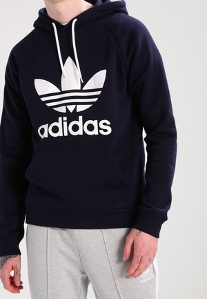 Osoba v tmavě modré mikině Adidas s bílým logem a světle šedých teplákoch Adidas, stojící před jednoduchým světlým pozadím.