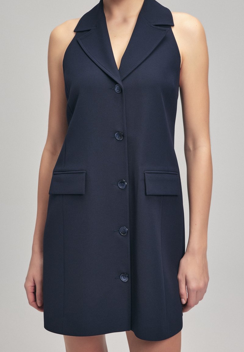 Robe bleu marine sans manches à boutons, avec un large col et deux poches à rabat à l'avant, portée par une personne avec les bras détendus sur les côtés.
