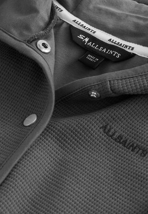 Harmaa tekstuurilla varustettu polo, jossa on kauluspääntie, nepparit ja brodeerattu "ALLSAINTS" -logo rinnassa. Hoito-ohjeet mukana.