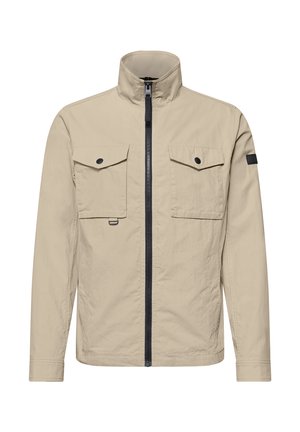 Veste beige légère avec fermeture éclair frontale, deux poches poitrine à rabat avec boutons noirs, manches longues et col court montant.