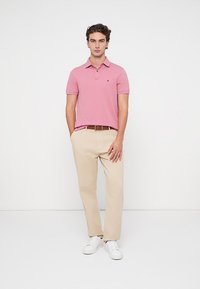 Rožinė polo marškinėliai su apykakle ir išsiuvinėtu logotipu, derintas su smėlinėmis kelnėmis ir baltais sportbačiais. Paprastas dizainas su laisvu siluetu.