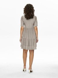 Robe beige à motifs avec des manches courtes bouffantes, un design en étages et des détails en dentelle, associée à des chaussures blanches à talons.
