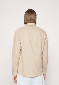 Beige overhemd met lange mouwen en een kraag. Gemaakt van een gestructureerde stof, voorzien van manchetten met knopen en een afgeronde zoom. Draagt bij lichte blauwe jeans.