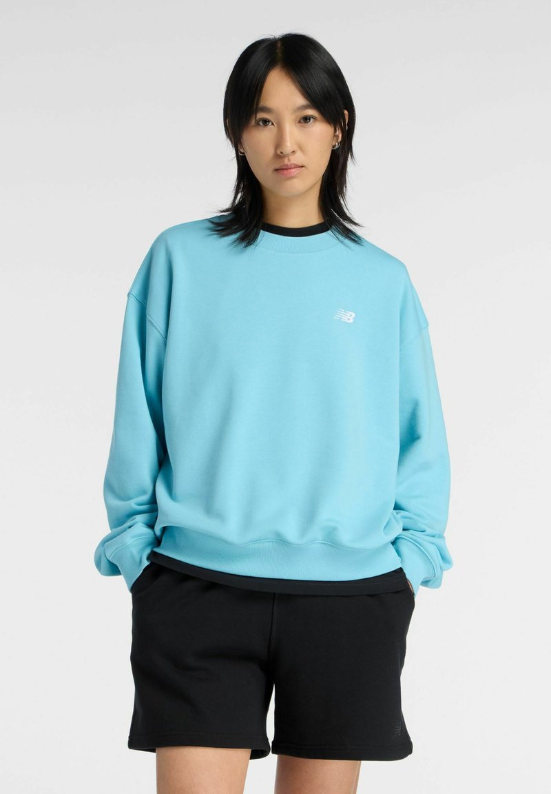 Jeune personne aux cheveux noirs portant un sweat-shirt bleu clair et un short noir, debout les mains dans les poches devant un fond uni.