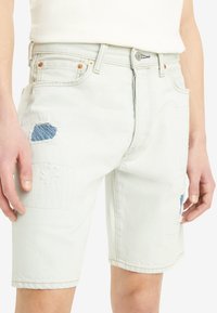 Shorts de mezclilla azul claro con parches desgastados, cinco bolsillos y accesorios de cobre. Presenta dobladillos enrollados y detalles de costura sutiles.