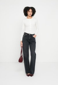 Άσπρο πουκάμισο με μακριά μανίκια και κουμπιά, σε συνδυασμό με ψηλόμεσο μαύρο τζιν παντελόνι φαρδιάς γραμμής· το μοντέλο κρατά μια μικρή μπορντό τσάντα.