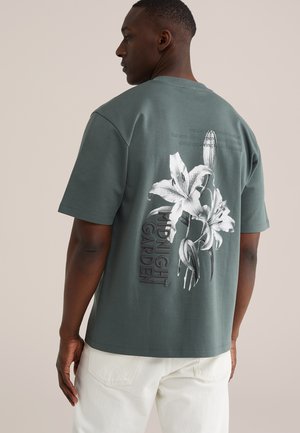 Muž v tlmene zelenej tričku s bielou ilustráciou ľalie a textom "MIDNIGHT GARDEN" na chrbte, v kombinácii s bielymi nohavicami.