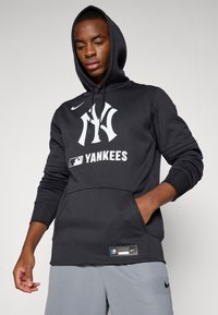 Juodas gobtuvėlis su Nike logotipu, didelė balta New York Yankees grafika, kengūros kišenė ir MLB etiketė ant krašto. Minkšta audinio tekstūra.