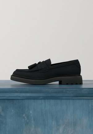 Mocassin en cuir noir avec glands, semelle épaisse en caoutchouc, talon plat, présenté sur une surface en bois bleue sur fond neutre.