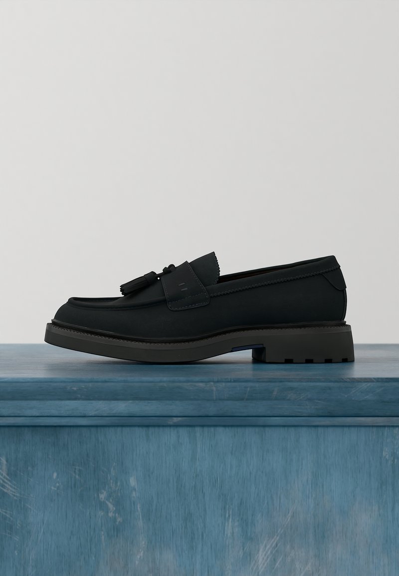 Mocassino nero in pelle con nappine, suola in gomma spessa, tacco basso, esposto su una superficie di legno blu su sfondo neutro.