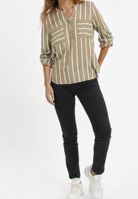 Femme portant un haut à rayures verticales tan et blanches avec des manches retroussées, un pantalon slim noir et des baskets blanches Adidas, se tenant devant un fond blanc.