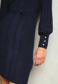 Marinblått stickat klänning med ribbat mönster, midjebälte och voluminösa ärmar som har tre silverknappar på ärmsluten.