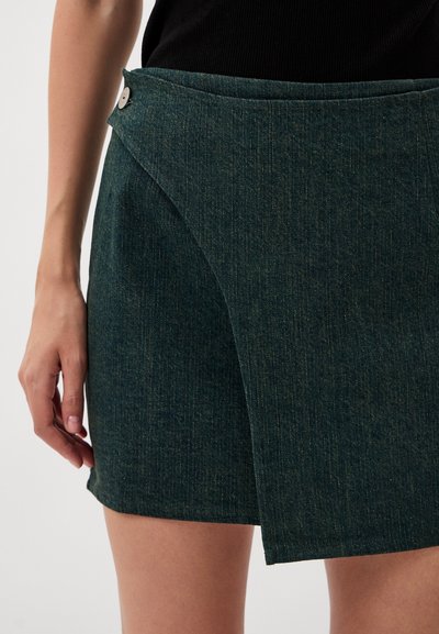 Green denim skirt featuring a wrap design, with a smooth texture, a front button closure, and minimal stitching details.