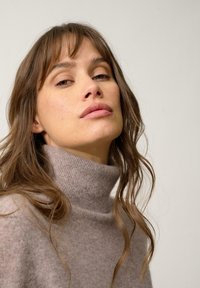 Maglione grigio chiaro a collo alto realizzato in morbido filato lavorato. Presenta una vestibilità comoda e una leggera lavorazione a costine sul collo. Sono visibili lunghi capelli mossi.