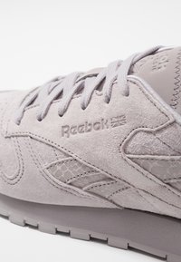 Ljusgrå Reebok-sneakers i mocka med texturerade accenter, utrustade med förstärkt häl, platta snören och en präglad logotyp på sidan.