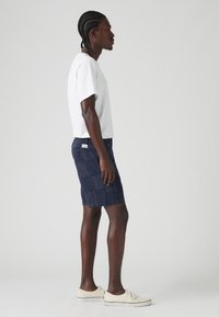 Tricou alb cu mânecă scurtă, pantaloni șort navy cu un design texturat și adidași deschiși. Prezentat în profil lateral pe un fundal neutru.