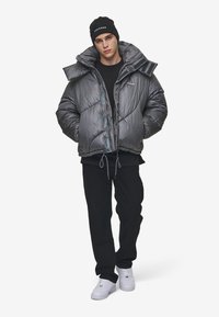 Pufferjack in metallic grijs met grote kraag, glanzende textuur, geribbelde accenten en een trekkoord onderaan. Draagt over een zwarte top en broek, met witte schoenen.