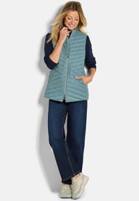 Frau lächelnd, trägt eine hellblaue Steppweste über einem marineblauen Pullover, dunkellblaue Jeans und weiße Sneakers, stehend vor einem weißen Hintergrund.