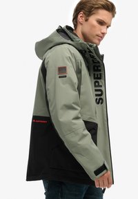 Grøngrå vandtæt jakke med sorte detaljer, stor hætte, frontlynlås, "SUPERDRY" logo og et bjerg-patch på ærmet.