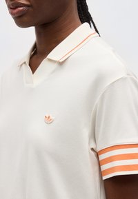 Κοντινό πλάνο ατόμου που φοράει λευκό πόλο πουκάμισο με πορτοκαλί ριγέ γιακά και μανσέτες στα μανίκια, καθώς και μικρό πορτοκαλί λογότυπο στο στήθος.