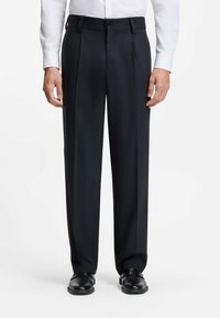 Zwarte geplooide broek van glad stof, met een knoopsluiting en zijzakken, gestyled met gepolijste zwarte schoenen.