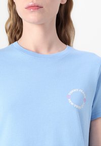 Šviesiai mėlynas medvilninis marškinėlis su apvaliu logotipo spaudiniu baltos ir rožinės spalvos, kuriame parašyta "TOMMY JEANS NEW YORK CITY" kairiajame krūtinės kampe. Trumpi rankoviai.