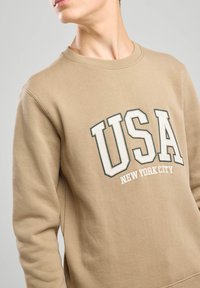 Beige sweatshirt with round neckline, featuring "USA" in bold white and green lettering and "NEW YORK CITY" below in smaller text.