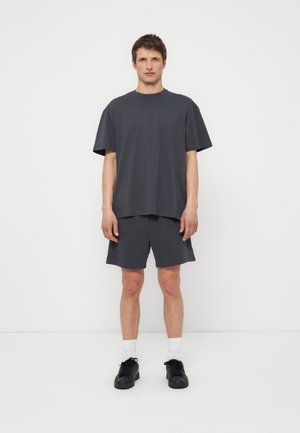 Férfi áll egy fehér fal előtt, sötétszürke, bő méretű pólót, hozzá illő rövidnadrágot, fehér zoknit és fekete tornacipőt visel, előre néz, karjai lazán lógnak.