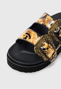 Crne natikače s tri ukrašena zlatno-crna remena; imaju ukrasnu metalnu opremu i teksturiranu potplatu.