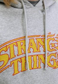 Haut à capuche gris avec le texte "STRANGE THINGS" imprimé en gras jaune avec des accents rouges. Il est doté d'une capuche à cordon et d'une texture douce.