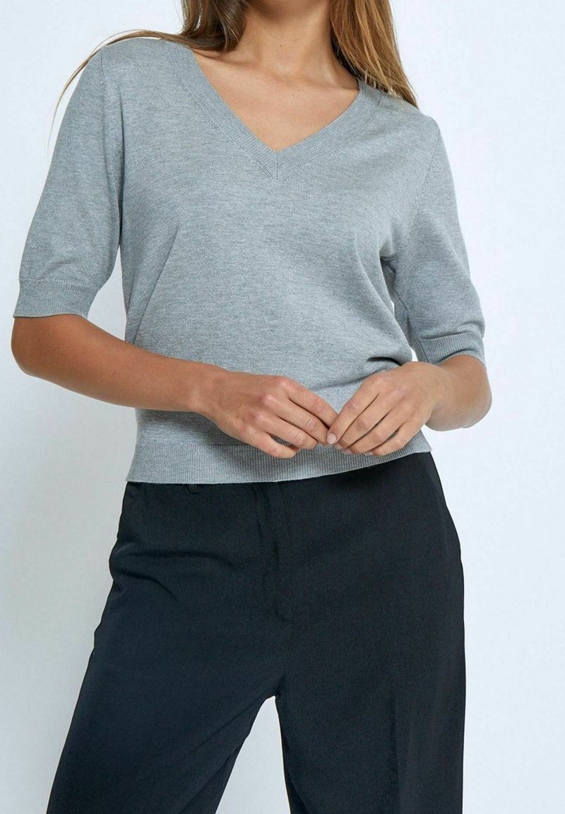 Femme portant un pull gris clair à manches courtes en V et un pantalon noir taille haute, mains jointes librement à la taille.