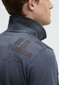 Grey, short-sleeve shirt with a high collar. Features printed text and graphics in darker tones. Visible stitching and a textured collar.