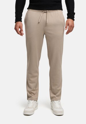 Homme portant un pantalon beige à cordon, des baskets blanches et un haut noir à manches longues, debout devant un fond blanc uni.