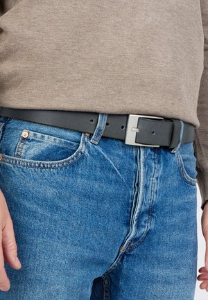 Sort læderbælte med en sølvfarvet rektangulær spænde, båret over blå denimjeans, parret med en lysebrun bluse med lange ærmer.