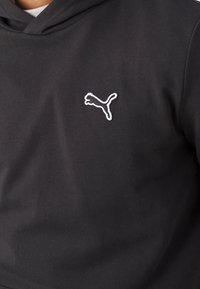 Sudadera con capucha negra hecha de tela suave con un bolsillo tipo canguro. Presenta un logo de Puma bordado en blanco en el pecho izquierdo. Diseño sencillo.