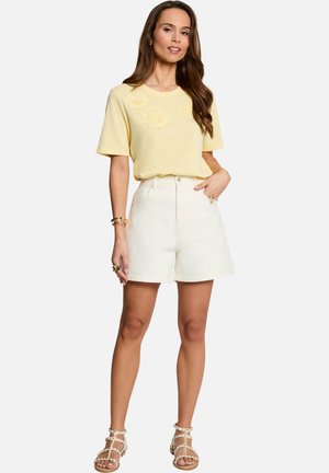 Femme portant un haut jaune à manches courtes avec broderies florales, un short blanc taille haute et des sandales blanches cloutées, debout avec une main dans la poche.