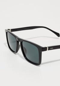 Óculos de sol retangulares pretos com lentes escuras. Armação de plástico espesso, com um detalhe prateado na haste com a marca "BOSS".
