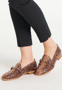 Lichte bruine loafers met slangenprint en een lage hak, voorzien van een glanzende gouden gesp als detail en een gestructureerde leren afwerking. Gelegen op een blote voet.